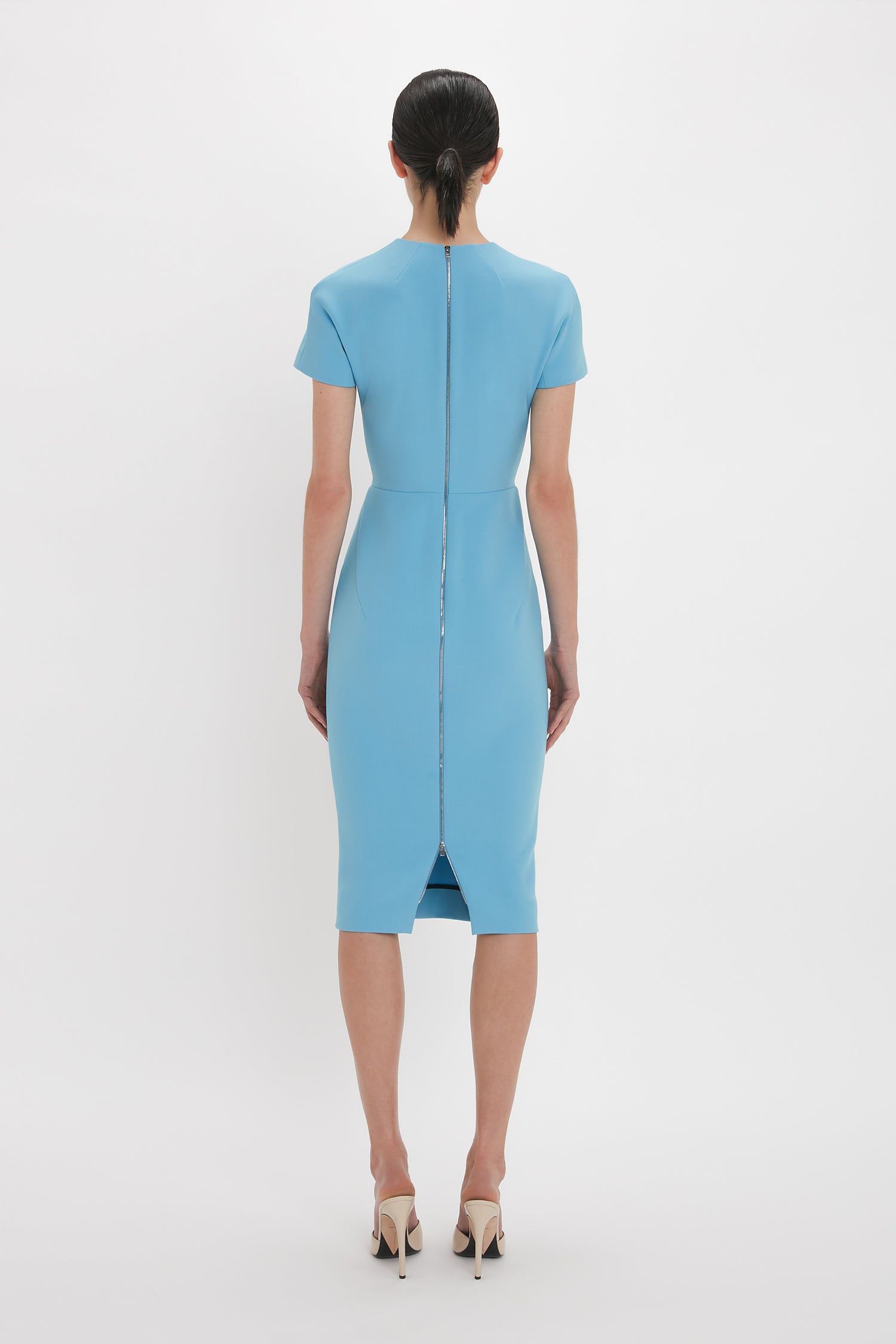 A woman is standing with her back to the camera, wearing the Victoria Beckham Exclusive Fitted T-Shirt Dress In Periwinkle Blue with short sleeves and a back zipper. She channels Victoria Beckham's timeless elegance, completed by her white high-heeled shoes.