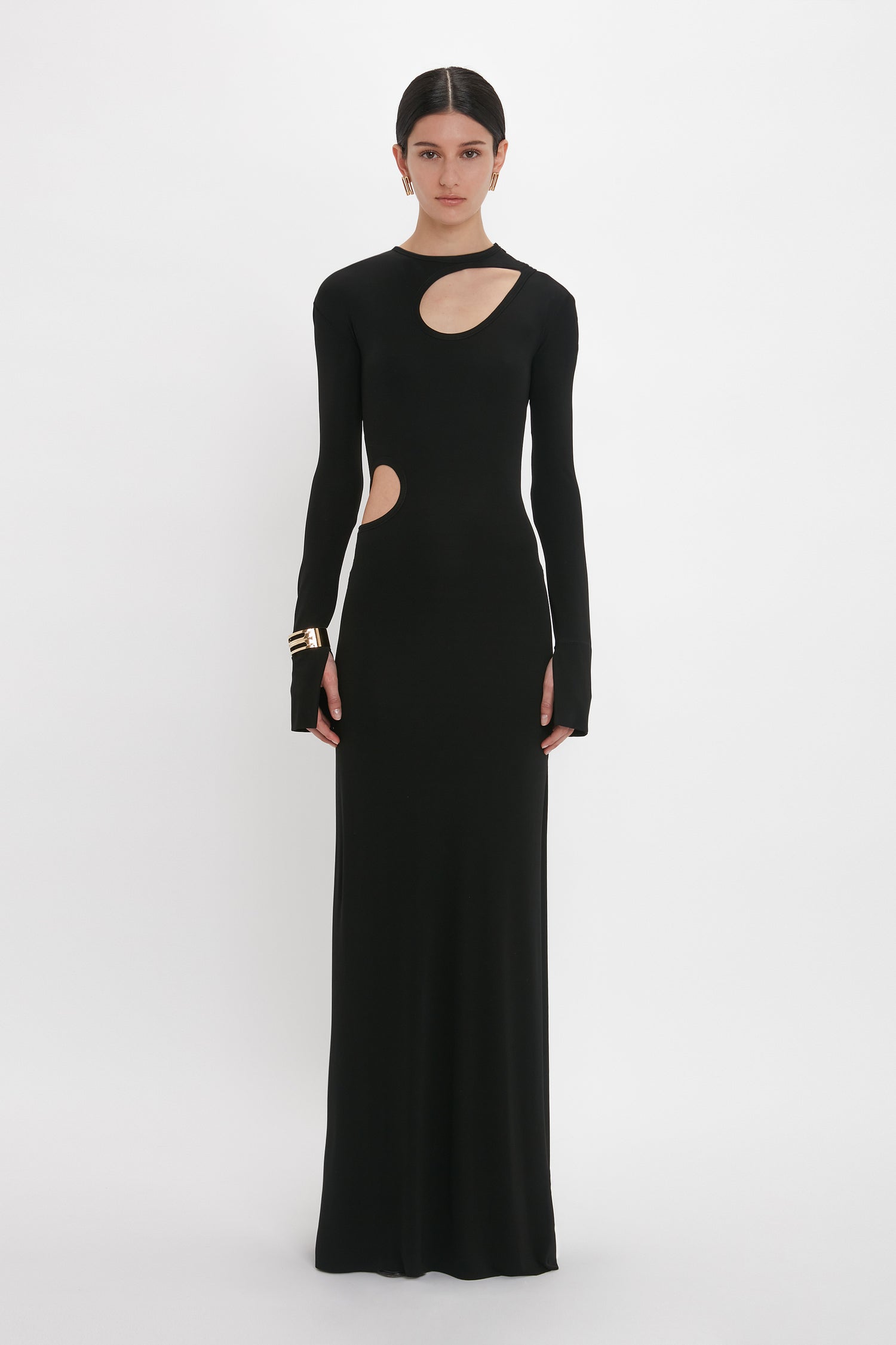 Person wearing a long, black, form-fitting evening gown with cut-out details on the chest and side, standing against a plain white background. The Cut-Out Jersey Floor-Length Dress In Black resembles a chic design from Victoria Beckham's collection.