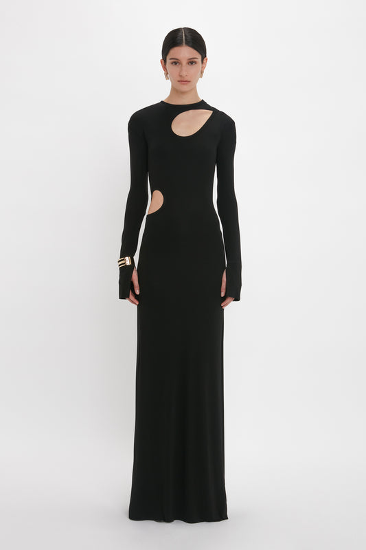 Person wearing a long, black, form-fitting evening gown with cut-out details on the chest and side, standing against a plain white background. The Cut-Out Jersey Floor-Length Dress In Black resembles a chic design from Victoria Beckham's collection.