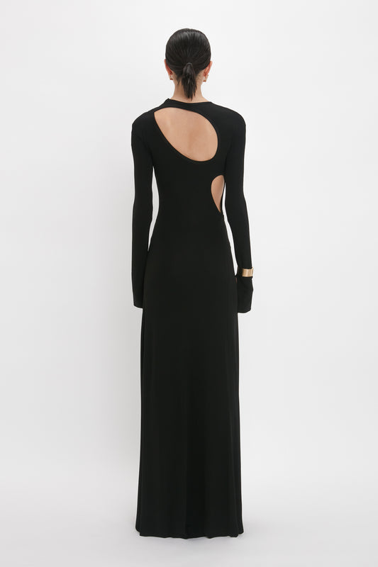 Person standing with their back facing the camera, wearing a long-sleeved Victoria Beckham Cut-Out Jersey Floor-Length Dress In Black with a circular cutout on the back and a gold bracelet on the left wrist.