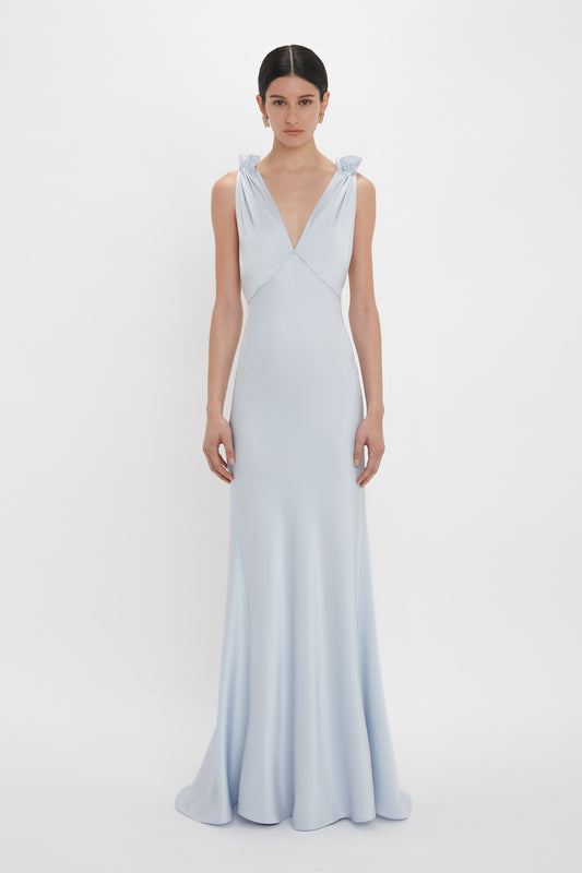 A person stands against a plain white background wearing an Exclusive Gathered Shoulder Cami Floor-Length Gown In Ice Blue by Victoria Beckham, with a deep V-neckline and bow details on the shoulders. The elegant ensemble appears to be crafted from luxurious crepe back satin.