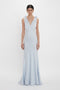 A person stands against a plain white background wearing an Exclusive Gathered Shoulder Cami Floor-Length Gown In Ice Blue by Victoria Beckham, with a deep V-neckline and bow details on the shoulders. The elegant ensemble appears to be crafted from luxurious crepe back satin.