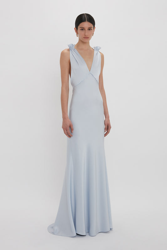 A woman stands in a Victoria Beckham Exclusive Gathered Shoulder Cami Floor-Length Gown In Ice Blue with tied shoulder straps and a deep V-neck, crafted from luxurious crepe back satin, against a plain white background.