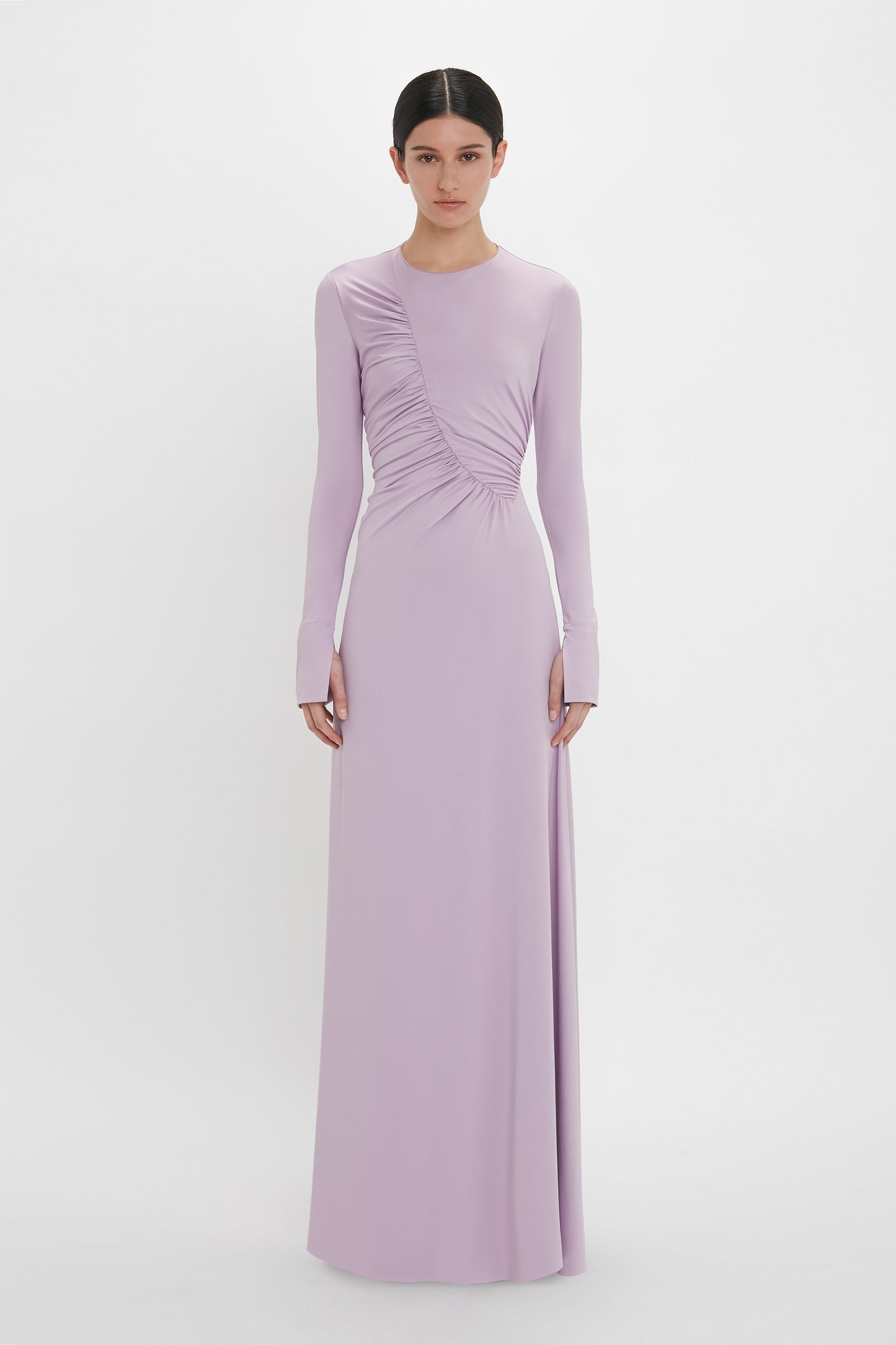 A person stands wearing a long-sleeved, floor-length light purple Ruched Detail Floor-Length Gown In Petunia by Victoria Beckham, crafted from stretch jersey fabric for understated glamour. The gathered detail on the right side adds a touch of elegance against the plain white background.