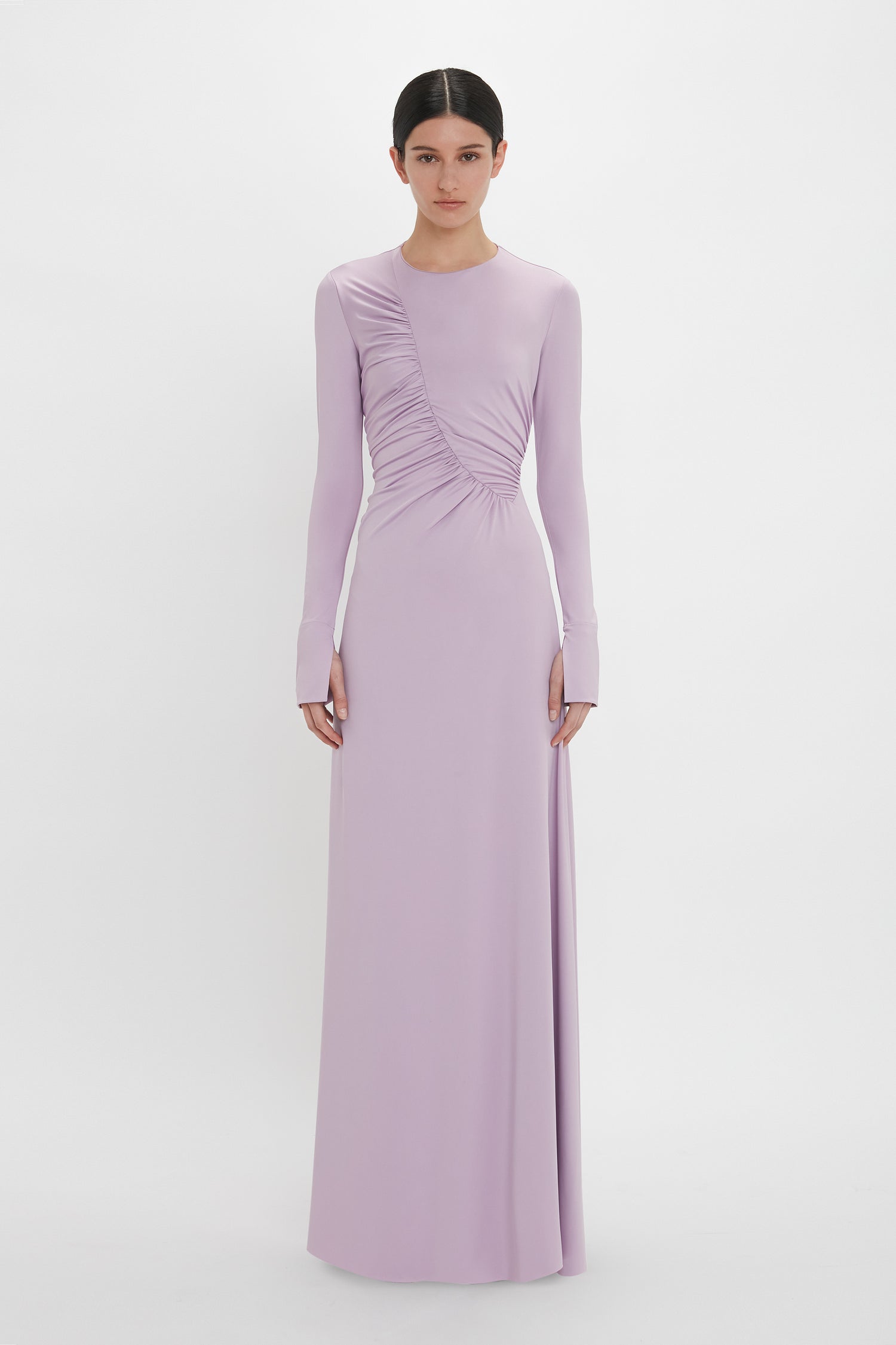 A person stands wearing a long-sleeved, floor-length light purple Ruched Detail Floor-Length Gown In Petunia by Victoria Beckham, crafted from stretch jersey fabric for understated glamour. The gathered detail on the right side adds a touch of elegance against the plain white background.