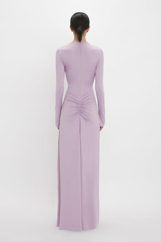 A person is standing with their back to the camera, wearing a long-sleeved, lavender-colored Ruched Detail Floor-Length Gown In Petunia from Victoria Beckham with gathered detailing at the lower back, embodying understated glamour.