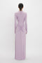 A person is standing with their back to the camera, wearing a long-sleeved, lavender-colored Ruched Detail Floor-Length Gown In Petunia from Victoria Beckham with gathered detailing at the lower back, embodying understated glamour.