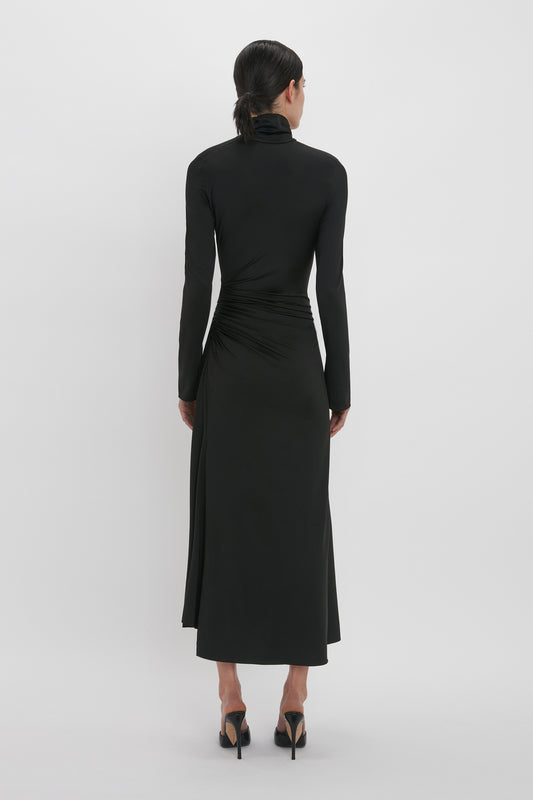 A woman in a Victoria Beckham high neckline, black long-sleeve dress and high heels viewed from behind against a plain background.