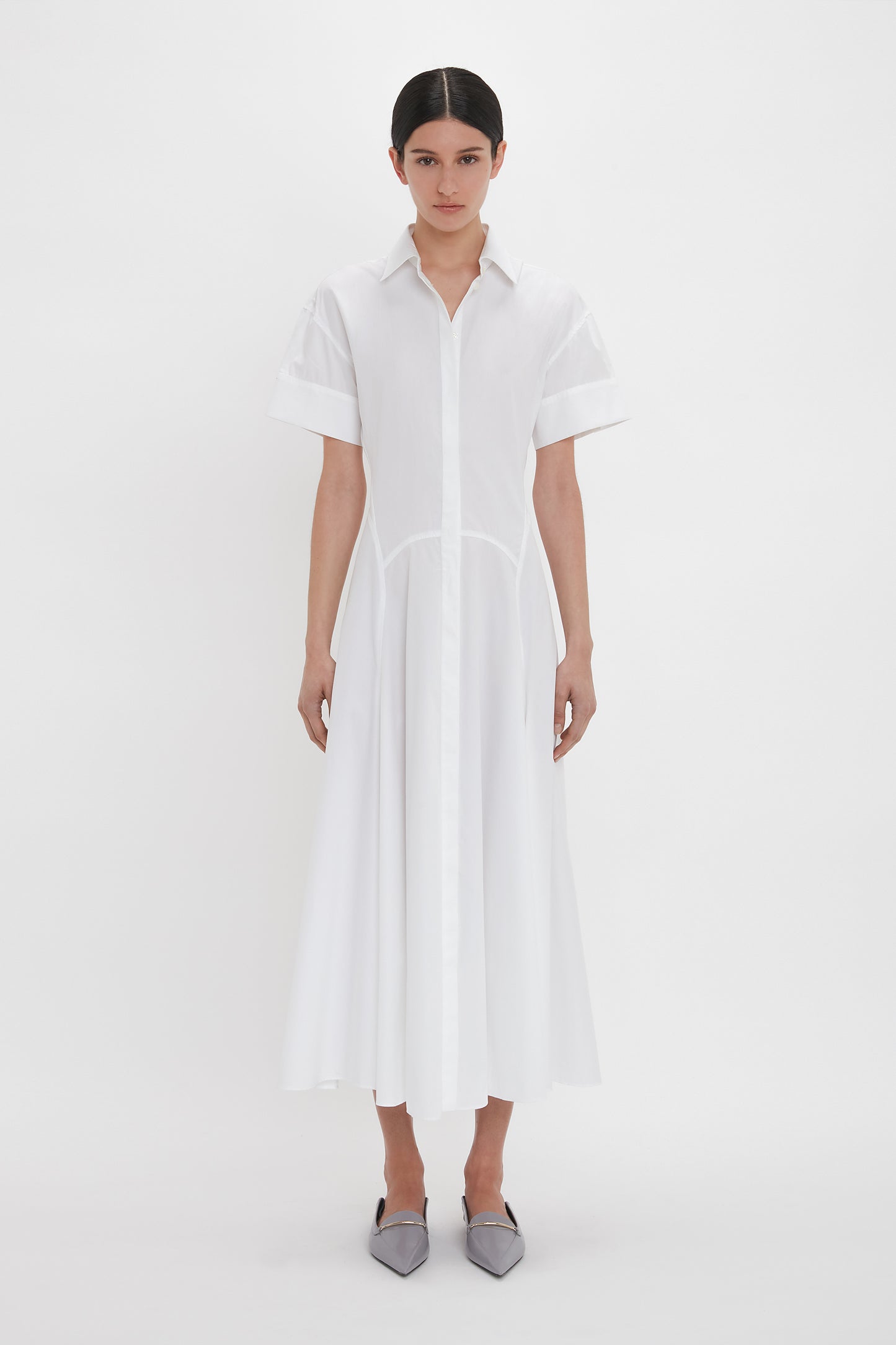 A person stands against a plain white background wearing the Victoria Beckham Panelled Shirt Dress In White and gray pointed shoes.