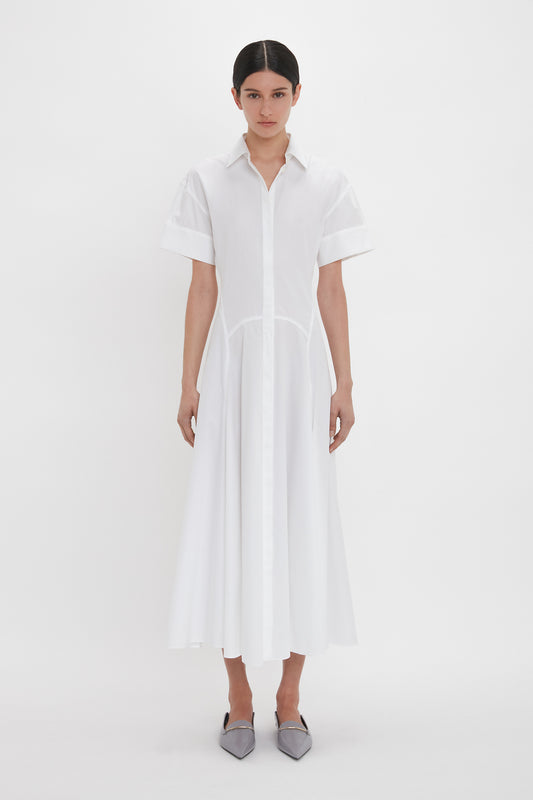 A person stands against a plain white background wearing the Victoria Beckham Panelled Shirt Dress In White and gray pointed shoes.