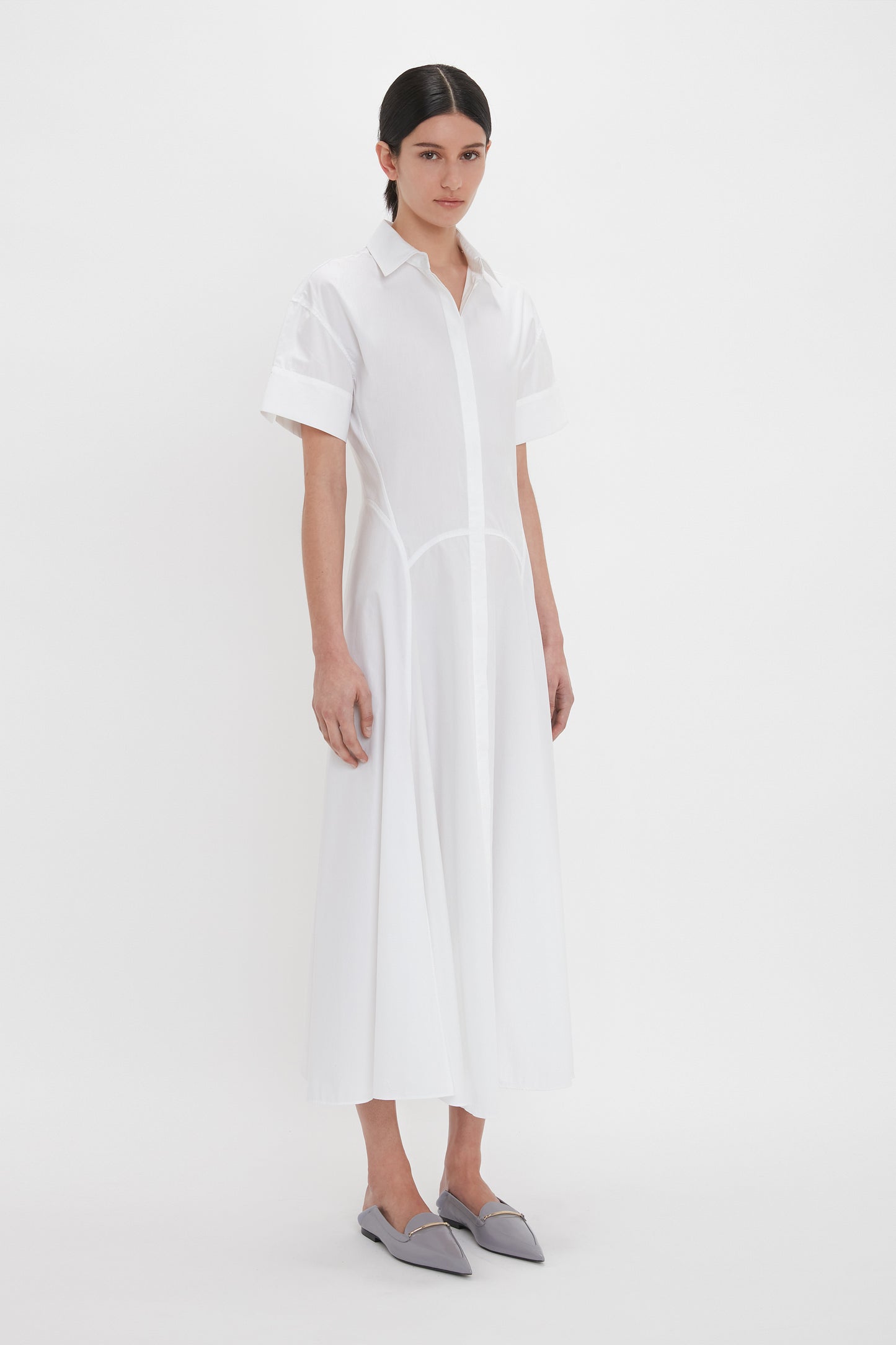 A person stands against a plain white background wearing the Panelled Shirt Dress In White by Victoria Beckham with short sleeves and grey pointed flats. The dress is crafted from organic cotton poplin, adding an eco-friendly touch to its crisp, elegant design.