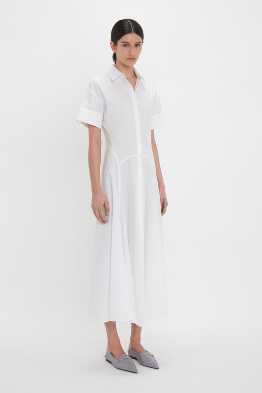 A person stands against a plain white background wearing the Panelled Shirt Dress In White by Victoria Beckham with short sleeves and grey pointed flats. The dress is crafted from organic cotton poplin, adding an eco-friendly touch to its crisp, elegant design.