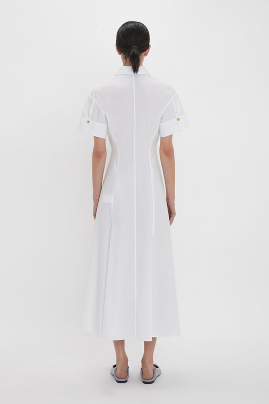 A woman stands facing away, wearing a Panelled Shirt Dress In White by Victoria Beckham, made from organic cotton poplin with gold button cuffs on the sleeves. She complements her look with white slip-on shoes.