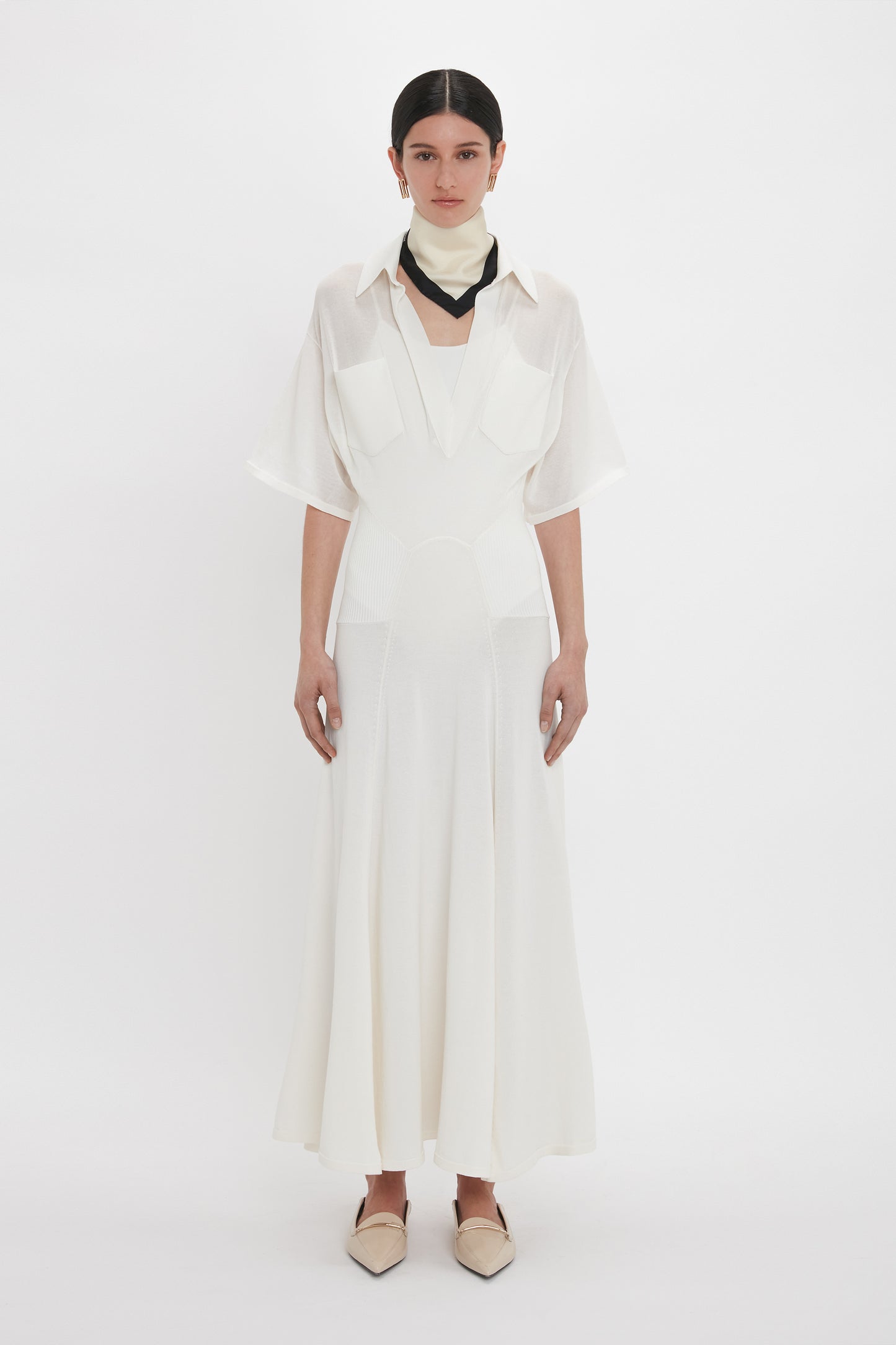 A person stands against a plain backdrop, exuding relaxed glamour in the Victoria Beckham Panelled Knit Dress In White with a collared shirt design and black neck accessory, paired with light-colored pointed shoes.