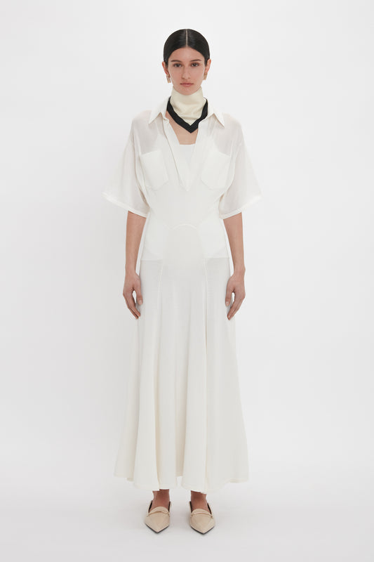 A person stands against a plain backdrop, exuding relaxed glamour in the Victoria Beckham Panelled Knit Dress In White with a collared shirt design and black neck accessory, paired with light-colored pointed shoes.