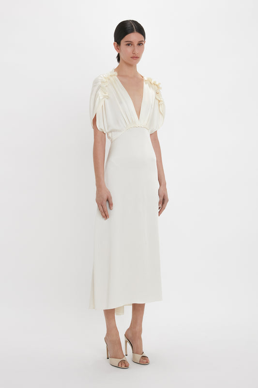A woman stands against a white background, wearing the Exclusive V-Neck Ruffle Midi Dress In Ivory by Victoria Beckham, featuring short, draped sleeves and tie detailing near the shoulders. She completes her look with open-toe heels.
