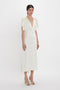 A woman stands against a white background, wearing the Exclusive V-Neck Ruffle Midi Dress In Ivory by Victoria Beckham, featuring short, draped sleeves and tie detailing near the shoulders. She completes her look with open-toe heels.