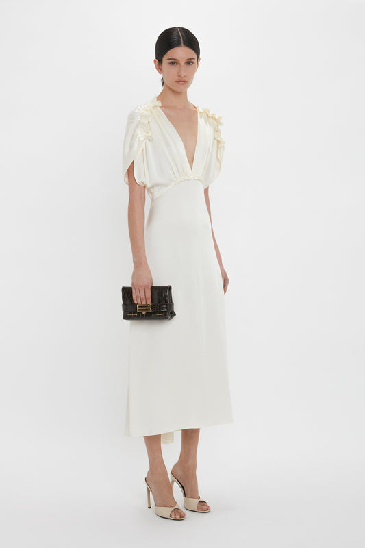 A woman stands against a white background wearing an elegant Exclusive V-Neck Ruffle Midi Dress In Ivory by Victoria Beckham, holding a black handbag, and wearing white heels.