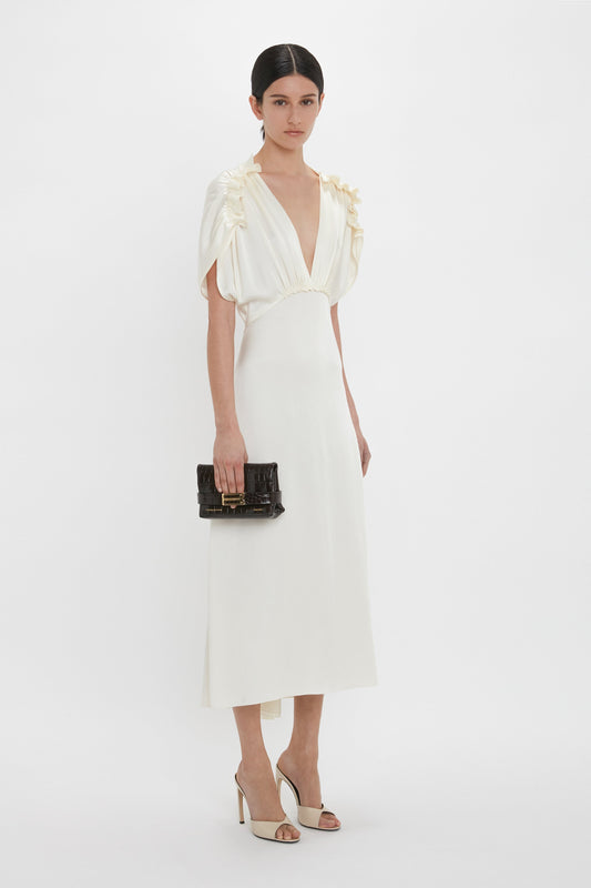 A woman stands against a plain background wearing a white dress with floral details on the shoulders, holding a Mini B Pouch Bag In Croc Effect Espresso Leather by Victoria Beckham, and dressed in beige high-heeled shoes.