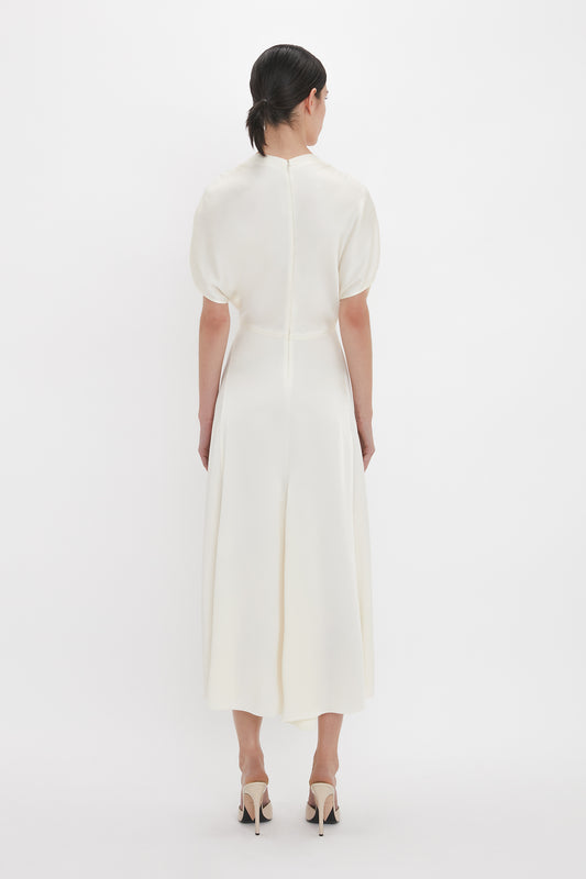 A person with dark hair, styled in a low bun, is wearing the Victoria Beckham Exclusive V-Neck Ruffle Midi Dress In Ivory and beige high heels, standing with their back facing the camera against a plain white background.