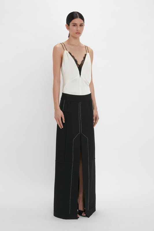 A woman stands against a plain background, wearing a white Lace Detail Cami Top In Harvest Ivory by Victoria Beckham with black lace trim, reminiscent of 1990s cami tops, paired with a long black crepe back satin skirt featuring a front slit.