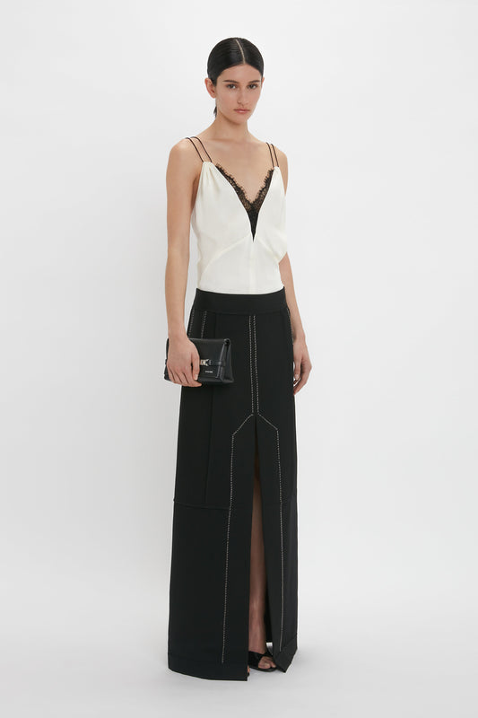 A woman stands in a white Lace Detail Cami Top In Harvest Ivory by Victoria Beckham with black lace detail and a long black skirt with a front slit, holding a black clutch bag.