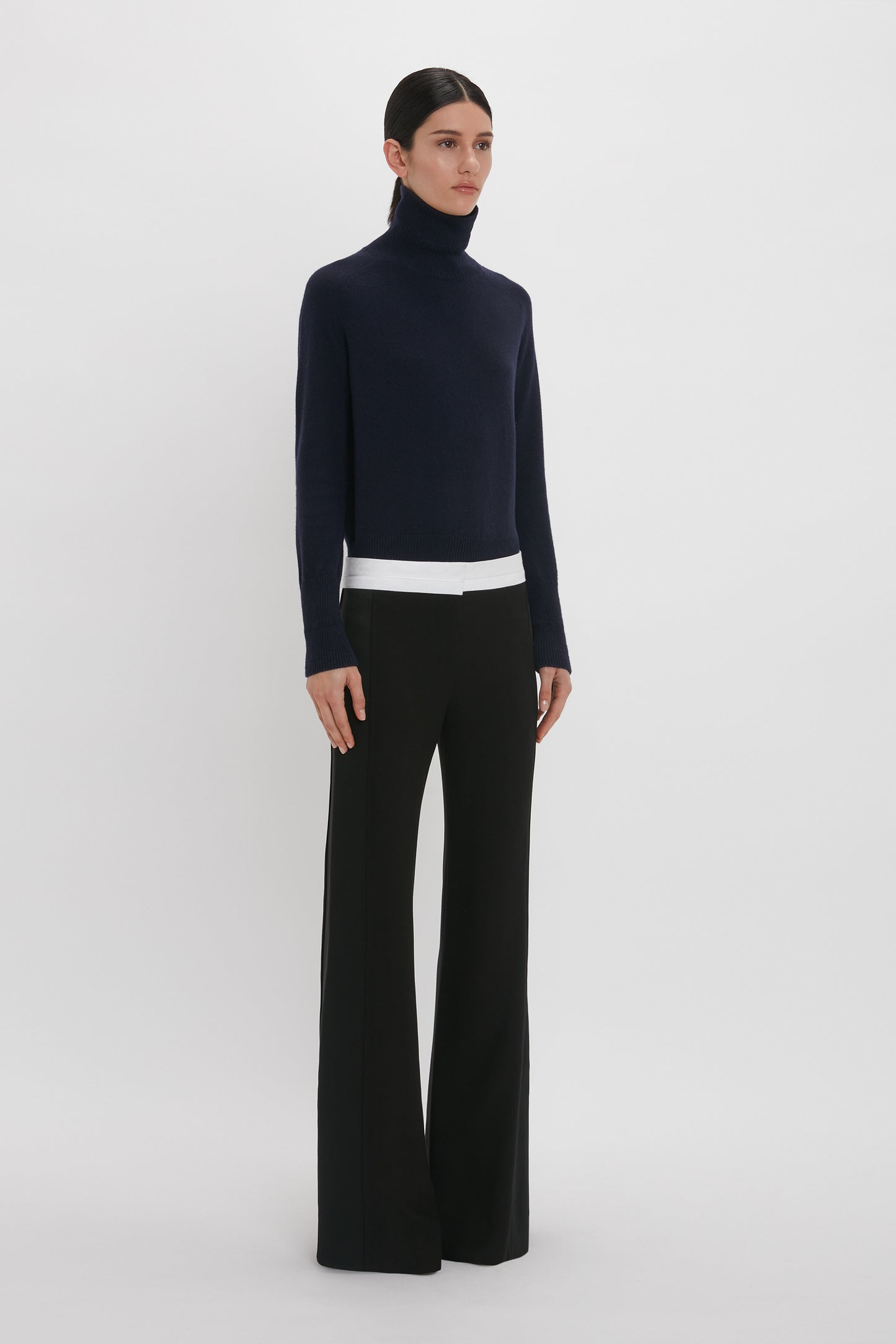A person stands against a plain background, wearing a black turtleneck sweater and Victoria Beckham's Side Panel Trouser In Black with a white waistband.