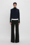 A person stands against a plain white background wearing a dark turtleneck sweater and Victoria Beckham's Side Panel Trouser In Black.