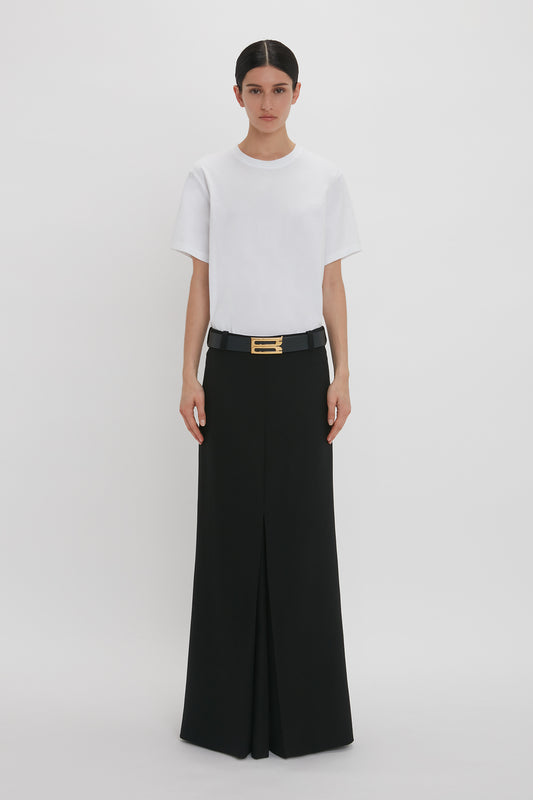 A woman wearing a white t-shirt and a Victoria Beckham black floor-length box pleat skirt with a gold buckle belt, standing against a plain white background.