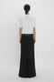 Woman viewed from behind wearing a white t-shirt and Floor-Length Box Pleat Skirt In Black by Victoria Beckham against a plain background.