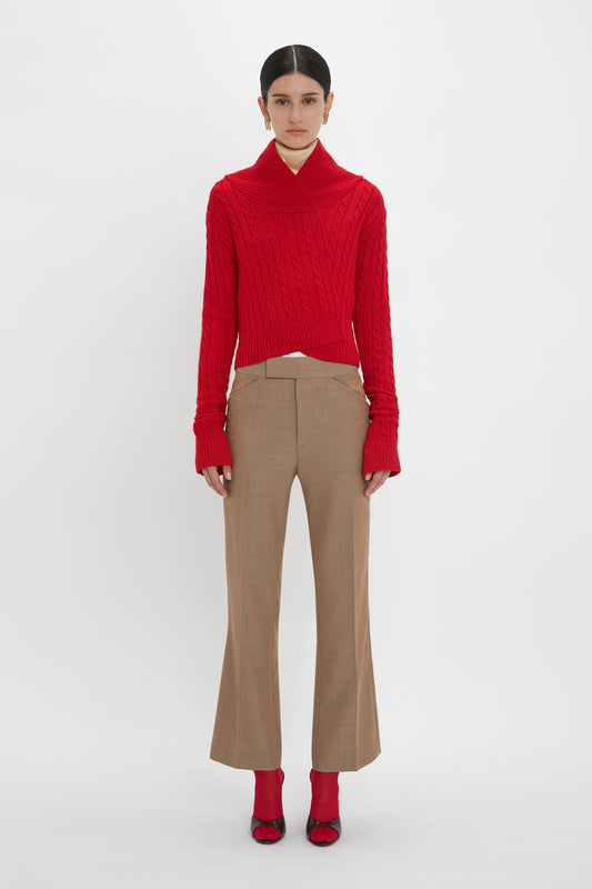 Person standing against a plain background, wearing a red Wrap Detail Jumper In Red by Victoria Beckham, tan high-waisted trousers, and red shoes.