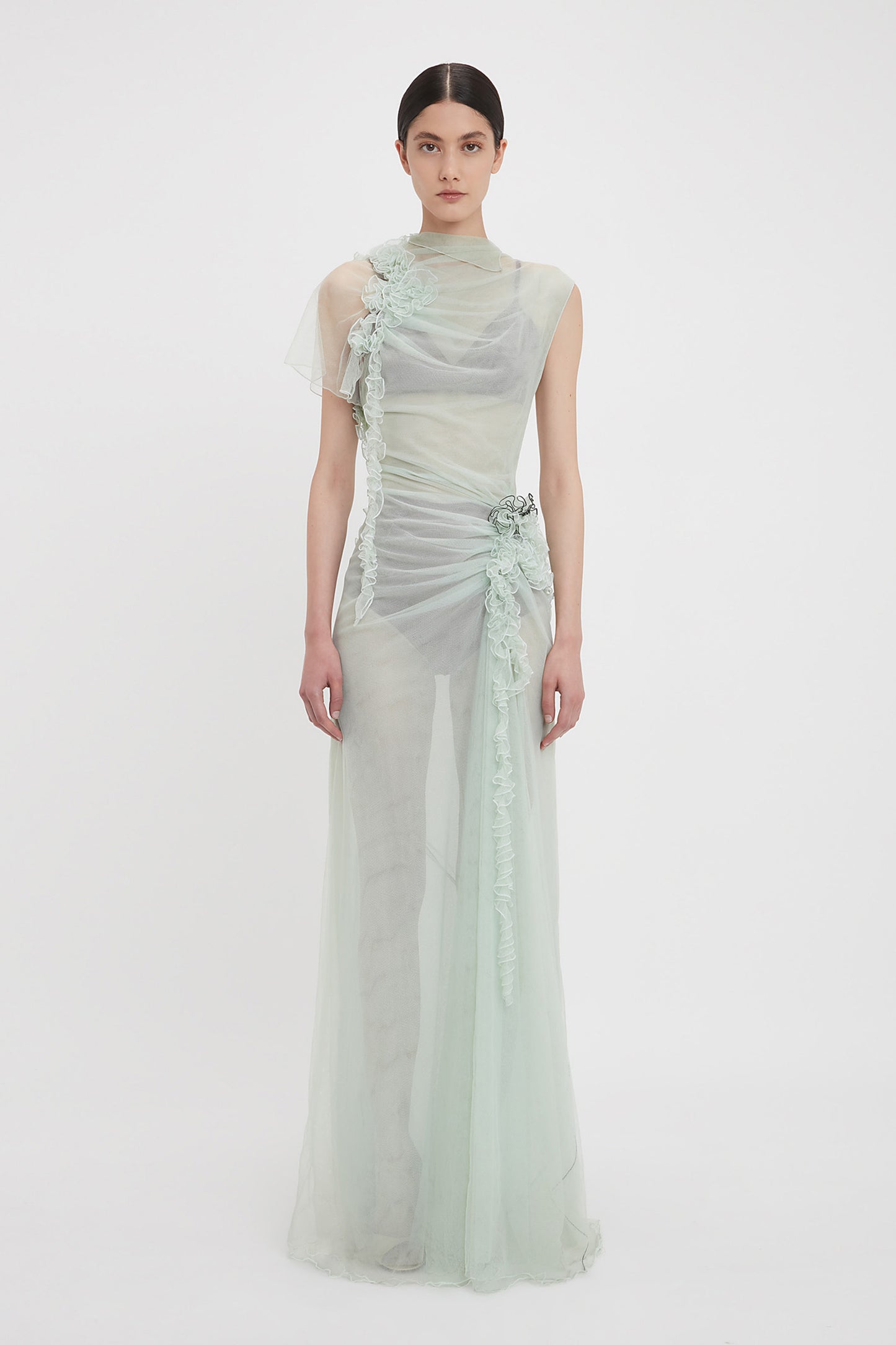 A woman stands against a white background wearing an ethereal, sea foam green tulle gown adorned with floral appliqués. The Gathered Tulle Detail Floor-Length Dress In Jade by Victoria Beckham has an asymmetrical design with a draped and flowing silhouette, embodying the elegance found at VictoriaBeckham.com.