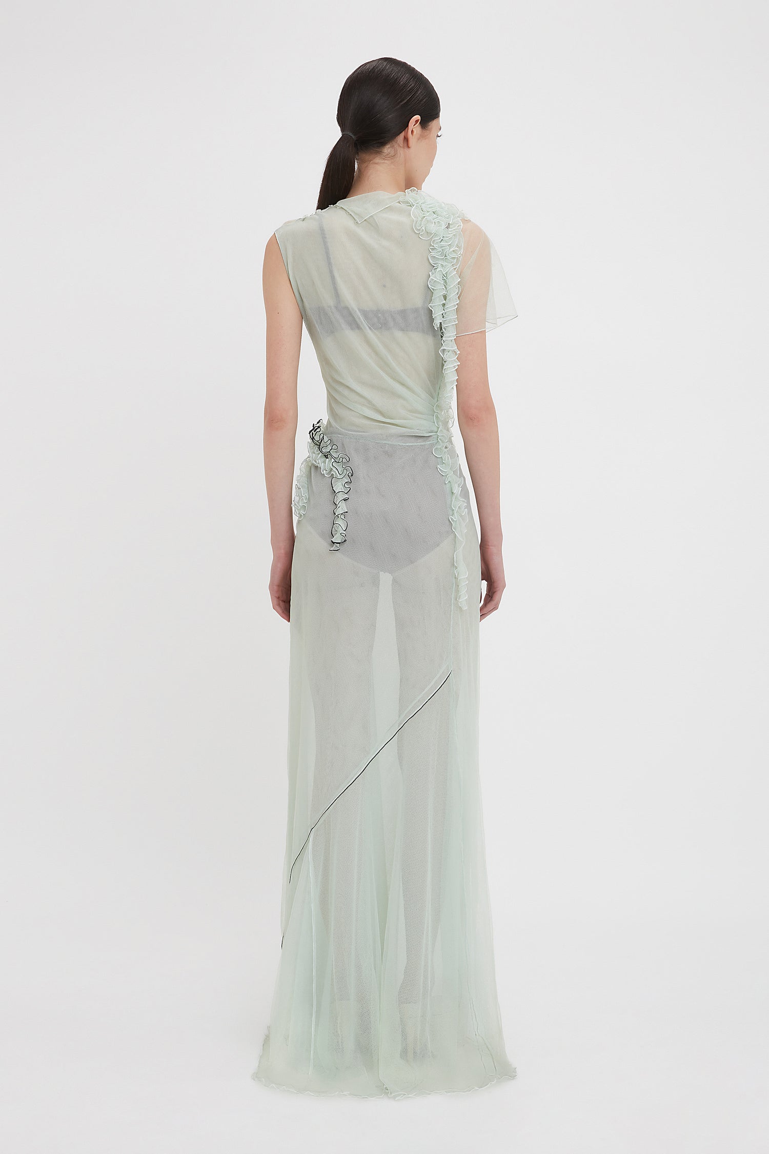 A person with long dark hair stands facing away, wearing a Gathered Tulle Detail Floor-Length Dress In Jade by Victoria Beckham against a plain white background. Discover more enchanting styles at VictoriaBeckham.com.