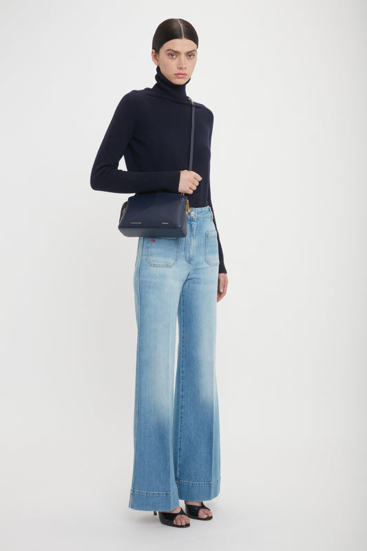 A person wearing a black turtleneck sweater, light blue high-waisted wide-leg jeans, and black open-toe heels, holding an Exclusive Victoria Crossbody Bag In Navy Leather by Victoria Beckham. The background is plain white.