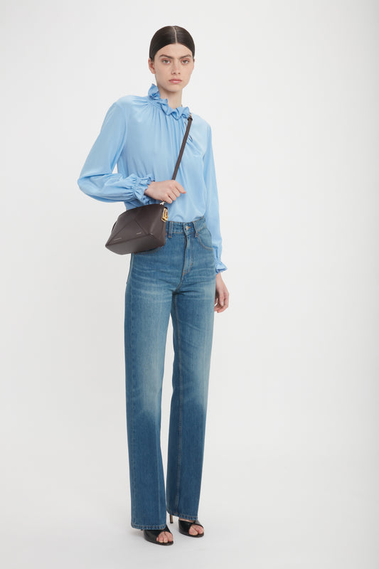 A person stands against a white background wearing a light blue blouse, high-waisted blue jeans, black sandals, and a brown Exclusive Victoria Crossbody Bag In Brown Leather by Victoria Beckham with an adjustable strap.