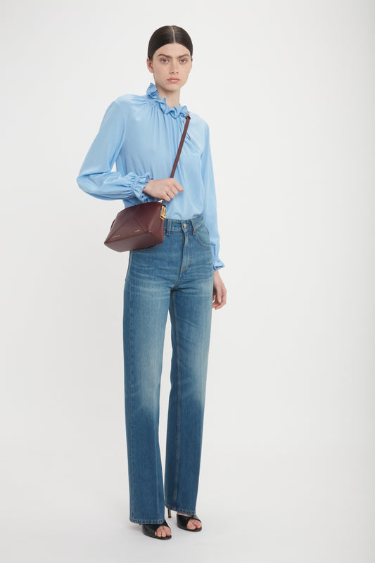 A person is standing against a plain background, wearing a light blue blouse, blue jeans, black open-toe heels, and carrying a Victoria Crossbody Bag In Burgundy Leather by Victoria Beckham.