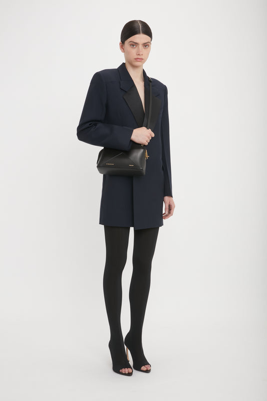 A person with a serious expression stands against a plain background, wearing a dark blazer, black leggings, and heels, carrying a black Victoria Beckham Victoria Crossbody In Black Leather made of calf leather with an adjustable strap.