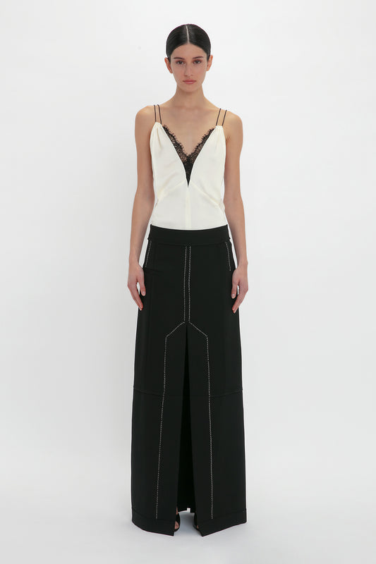 A person stands against a plain background wearing an elegant Lace Detail Cami Top In Harvest Ivory by Victoria Beckham and a long black skirt with white stitching.