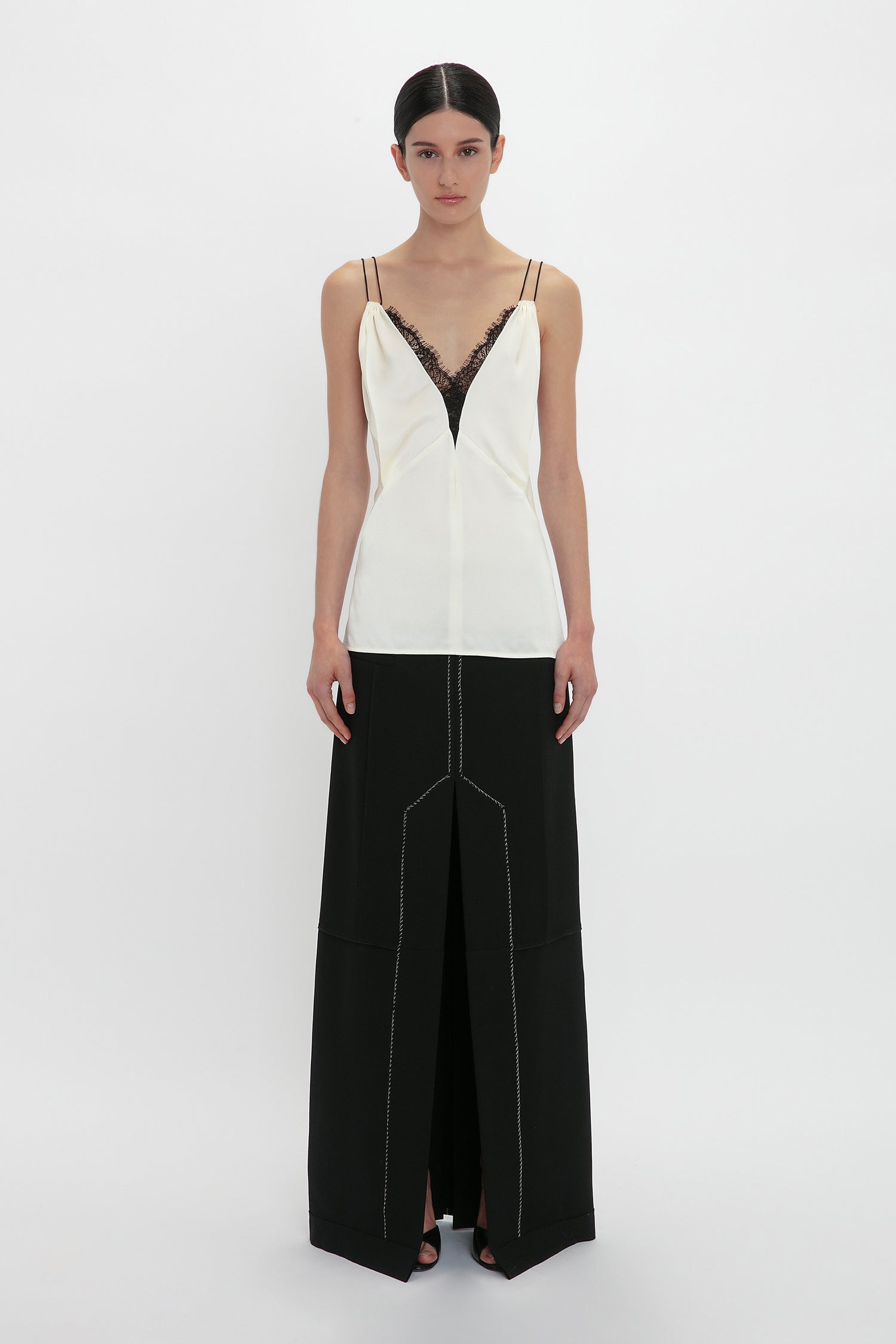 A person stands against a white background, wearing a Lace Detail Cami Top In Harvest Ivory by Victoria Beckham and a long black skirt with a front slit.