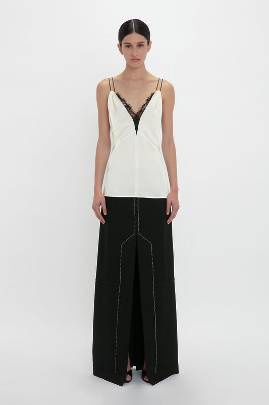 A person stands against a white background, wearing a Lace Detail Cami Top In Harvest Ivory by Victoria Beckham and a long black skirt with a front slit.