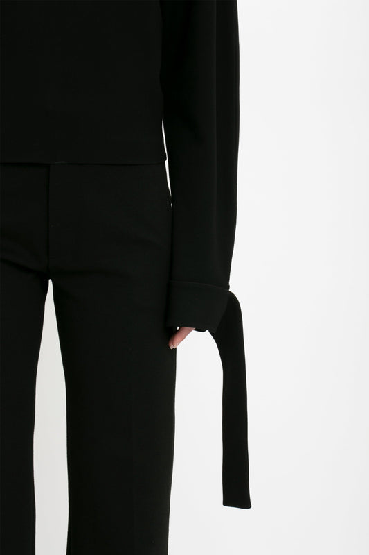 A person wearing a black Victoria Beckham Tie Sleeve Ponti Top In Black and matching black pants, with long ties hanging from the sleeves, stands against a plain white background, embodying chic off-duty dressing.