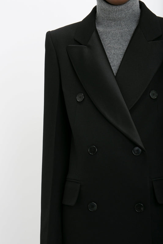 Person wearing a slim fit Victoria Beckham Double Breasted Tuxedo Coat in Black over a gray turtleneck sweater against a plain background, partially cropped to show the upper body.