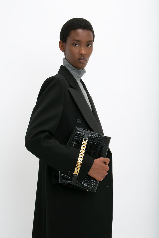 A person in a Double Breasted Tuxedo Coat in Black by Victoria Beckham and gray turtleneck holds a black and gold handbag against a plain white background.