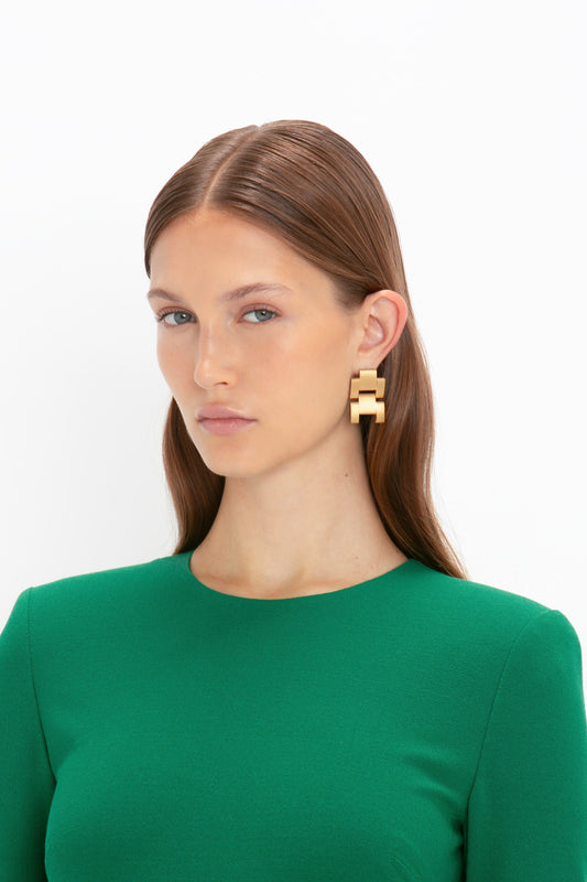 A person with long brown hair wearing a Victoria Beckham Long Sleeve T-Shirt Fitted Dress in Emerald made of bi-stretch crepe fabric, complemented by large gold geometric earrings, stands against a white background.