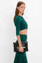 A person stands against a white background, wearing a teal green *VB Body Cropped Cardi In Lurex Green* by *Victoria Beckham* and skirt, showcasing a form-fitting silhouette, and holding a black clutch with a gold chain detail.