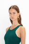 A woman with long brown hair wears a VB Body Sleeveless Dress In Lurex Green by Victoria Beckham and gold geometric earrings, posing against a white background. Her new-season wardrobe features a flattering flared silhouette, perfect for any occasion.