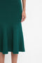 Close-up of a person wearing a teal VB Body Flared Skirt In Lurex Green by Victoria Beckham, standing against a white background. Only the lower body and right hand are visible.