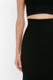 Close-up of a woman standing against a white background wearing a black crop top and high-waisted VB Body Scallop Trim Flared Skirt In Black from Victoria Beckham. Her left arm is visible and relaxed by her side.