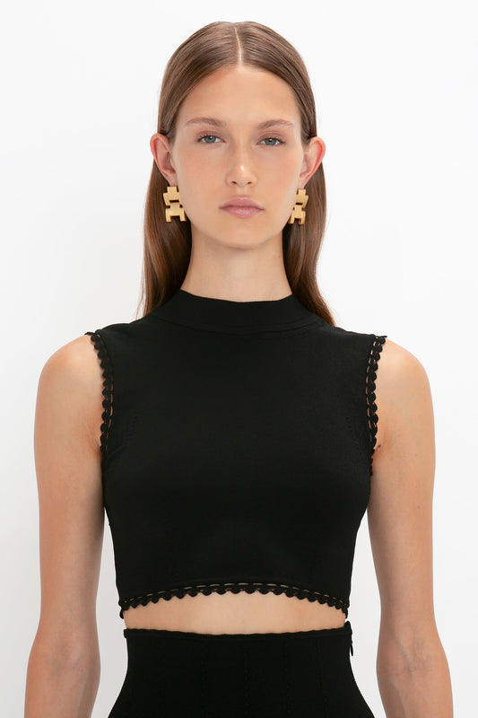 A person with long hair wearing a versatile, sleeveless black Victoria Beckham VB Body Scallop Trim Tank Top In Black and geometric earrings stands against a plain white background, looking directly at the camera.