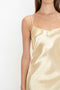 A woman wearing a shiny, cream-colored Exclusive Floor-Length Cami Dress In Gold by Victoria Beckham with spaghetti straps is shown from the chest down, evoking a 1990s-inspired elegance. The background is plain and white.